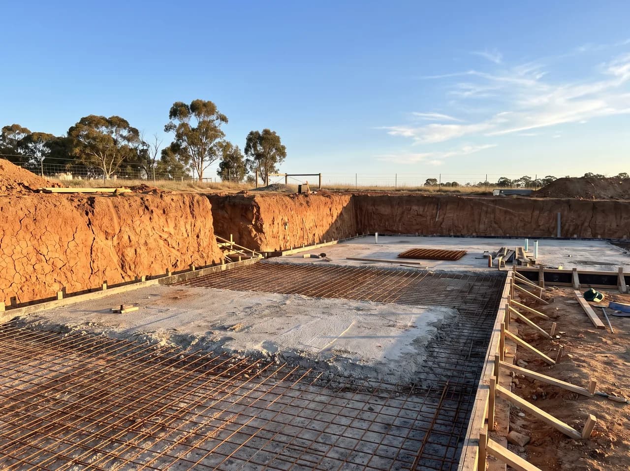 An excavated building block with exposed reactive clay and a partly poured slab in warm afternoon light