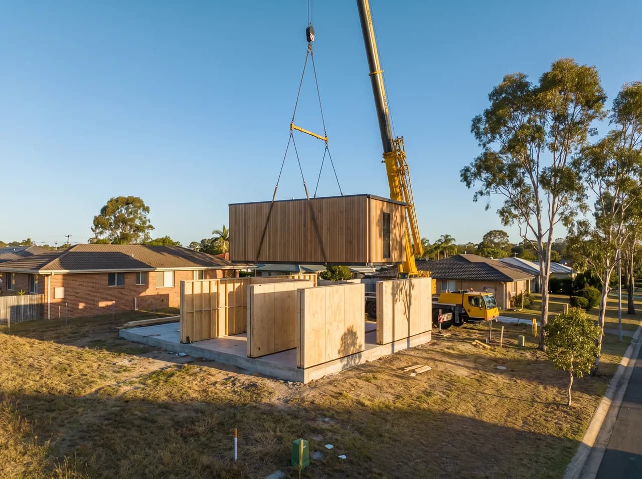 Flat-pack modular wall panels and a craned prefab module being positioned on a simple block in warm afternoon light